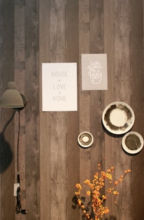 A wooden wall features two framed prints with motivational text, a modern wall lamp, and decorative ceramic plates. Bright orange branches provide a pop of color at the bottom.