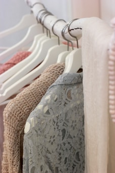 A close-up view of clothing hanging on a white wooden rack. The garments feature a variety of textures, including knit and lace, in soft and neutral colors like beige, pink, and blue. The hangers are uniformly white.