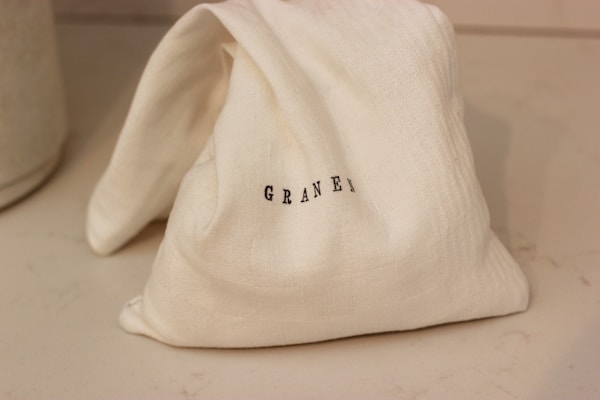A small, folded cloth bag with the word 'GRAINGER' printed in black letters. It rests on a smooth, light-colored surface with shadows indicating soft lighting.