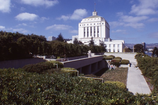 A large white government building with classical architectural features stands prominently against a background of greenery and a partly cloudy blue sky. In the foreground, manicured bushes and a concrete walkway lead towards the building.