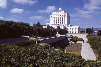 A large white government building with classical architectural features stands prominently against a background of greenery and a partly cloudy blue sky. In the foreground, manicured bushes and a concrete walkway lead towards the building.