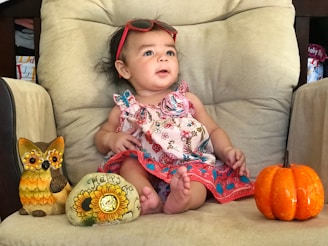 A baby is seated on a beige armchair wearing a colorful floral dress and red sunglasses on the head. Surrounding the baby are decorative items including a ceramic owl, a painted stone with a sunflower and 'Happy Fall' written on it, and a small orange pumpkin.