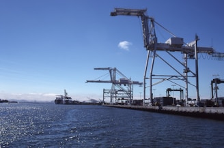 A port scene with several large cranes on a dock, extending over the water. The sky is clear, and the sun glistens on the surface of the water. Shipping containers and port equipment are visible along the dock.