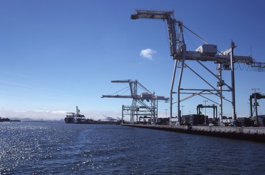 A port scene with several large cranes on a dock, extending over the water. The sky is clear, and the sun glistens on the surface of the water. Shipping containers and port equipment are visible along the dock.