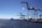 A port scene with several large cranes on a dock, extending over the water. The sky is clear, and the sun glistens on the surface of the water. Shipping containers and port equipment are visible along the dock.