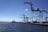 A port scene with several large cranes on a dock, extending over the water. The sky is clear, and the sun glistens on the surface of the water. Shipping containers and port equipment are visible along the dock.
