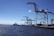 A port scene with several large cranes on a dock, extending over the water. The sky is clear, and the sun glistens on the surface of the water. Shipping containers and port equipment are visible along the dock.