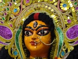 A detailed close-up of a vibrant and intricately designed religious idol featuring ornate decorations and jewelry. The face is painted with bold black lines emphasizing the eyes and features a central bindi. The headgear is elaborately adorned with colorful floral patterns and sequins in gold, purple, blue, and green hues.