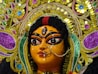 A detailed close-up of a vibrant and intricately designed religious idol featuring ornate decorations and jewelry. The face is painted with bold black lines emphasizing the eyes and features a central bindi. The headgear is elaborately adorned with colorful floral patterns and sequins in gold, purple, blue, and green hues.