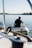 A close-up view of a boat deck with stainless steel fittings and a sheet pulley in focus. A person, slightly out of focus, is seated in a small inflatable boat on the water. The calm water and blurred horizon create a serene backdrop.