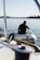 A close-up view of a boat deck with stainless steel fittings and a sheet pulley in focus. A person, slightly out of focus, is seated in a small inflatable boat on the water. The calm water and blurred horizon create a serene backdrop.