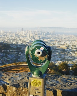 A green public viewer telescope is positioned on a stone ledge overlooking a panoramic view of a sprawling cityscape with skyscrapers in the distance. The landscape is bathed in natural light, showcasing a blend of urban and natural scenery.