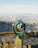 A green public viewer telescope is positioned on a stone ledge overlooking a panoramic view of a sprawling cityscape with skyscrapers in the distance. The landscape is bathed in natural light, showcasing a blend of urban and natural scenery.