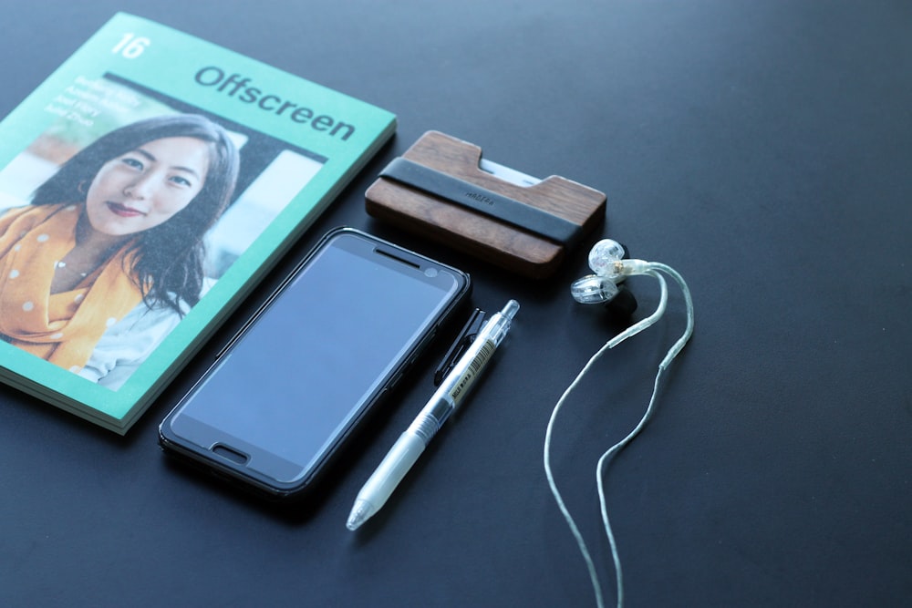 black Android smartphone beside white earbuds, white click pen, and Offscreen book