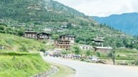 A scenic village set against a backdrop of lush green hills and dense forests. Traditional wooden houses with distinct architectural styles are scattered across the hillside. A winding road leads through the village, with a few vehicles and people visible near the roadside. Brightly colored umbrellas add contrast to the greenery.