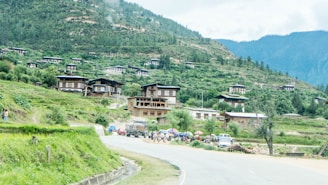 A scenic village set against a backdrop of lush green hills and dense forests. Traditional wooden houses with distinct architectural styles are scattered across the hillside. A winding road leads through the village, with a few vehicles and people visible near the roadside. Brightly colored umbrellas add contrast to the greenery.