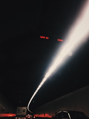A dark tunnel illuminated by a bright, narrow line of lights along the ceiling. Several vehicles are visible in the tunnel, including cars and a truck. The red text 'TURN ON RADIO' is visible at the top.
