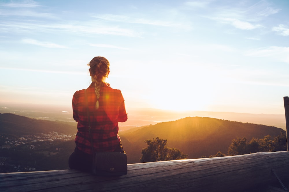 person sitting on wood fence facing mountain under cloudy sky