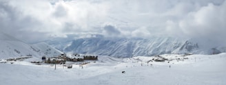 A vast, snow-covered landscape with a small village of chalets located in the middle. The village is surrounded by ski slopes with several people skiing and moving up ski lifts. The background features majestic, snow-capped mountains under a cloudy sky. The overall scene gives a sense of stillness and cold weather.