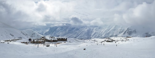 A vast, snow-covered landscape with a small village of chalets located in the middle. The village is surrounded by ski slopes with several people skiing and moving up ski lifts. The background features majestic, snow-capped mountains under a cloudy sky. The overall scene gives a sense of stillness and cold weather.