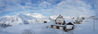 A snowy mountain landscape with traditional alpine-style wooden houses covered in snow. The sky is partly cloudy with patches of blue visible. Snow blankets the ground and the surrounding mountains, creating a serene, winter atmosphere.