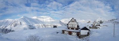 A snowy mountain landscape with traditional alpine-style wooden houses covered in snow. The sky is partly cloudy with patches of blue visible. Snow blankets the ground and the surrounding mountains, creating a serene, winter atmosphere.