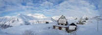 A snowy mountain landscape with traditional alpine-style wooden houses covered in snow. The sky is partly cloudy with patches of blue visible. Snow blankets the ground and the surrounding mountains, creating a serene, winter atmosphere.