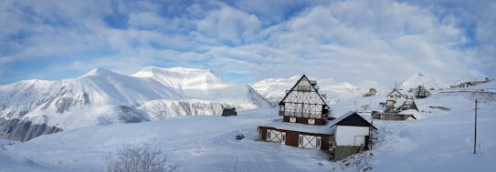 A snowy mountain landscape with traditional alpine-style wooden houses covered in snow. The sky is partly cloudy with patches of blue visible. Snow blankets the ground and the surrounding mountains, creating a serene, winter atmosphere.