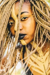 A close-up of a person with braided hair and artistic face paint, featuring gold and black colors. The face paint includes a bold stripe and eye makeup, and the person has dark lipstick and facial piercings.