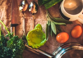 A composition of kitchen items and ingredients arranged on a cloth surface. There is a knife and two spoons with reflective surfaces, fresh kale leaves, sage sprigs, two brown eggs, a white mug with a black design, and a green figurine resembling a relaxed Buddha. A shiny metallic pan is also visible.