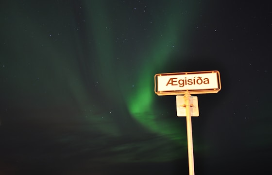 A street sign with the name 'Ægisíða' stands against a backdrop of a vivid green aurora in the night sky, speckled with stars. The sign is illuminated, contrasting with the dark and ethereal scenery.