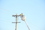 A worker in a bucket lift is performing maintenance on a utility pole, wearing protective gear including a hard hat. The utility pole has multiple wires attached, and the scene is set against a clear blue sky.
