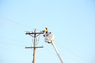 A worker in a bucket lift is performing maintenance on a utility pole, wearing protective gear including a hard hat. The utility pole has multiple wires attached, and the scene is set against a clear blue sky.