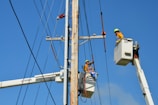 Two utility workers in safety gear are using bucket trucks to work on a power line. The sky is clear and blue, providing a backdrop to the scene. Both workers are elevated in their respective buckets, focusing on tasks related to the maintenance or repair of the electrical infrastructure.