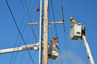 Two utility workers in safety gear are using bucket trucks to work on a power line. The sky is clear and blue, providing a backdrop to the scene. Both workers are elevated in their respective buckets, focusing on tasks related to the maintenance or repair of the electrical infrastructure.