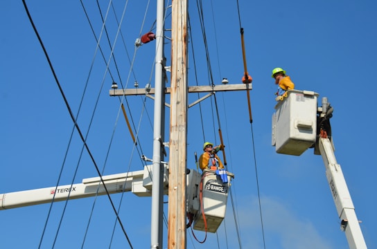 Two utility workers in safety gear are using bucket trucks to work on a power line. The sky is clear and blue, providing a backdrop to the scene. Both workers are elevated in their respective buckets, focusing on tasks related to the maintenance or repair of the electrical infrastructure.