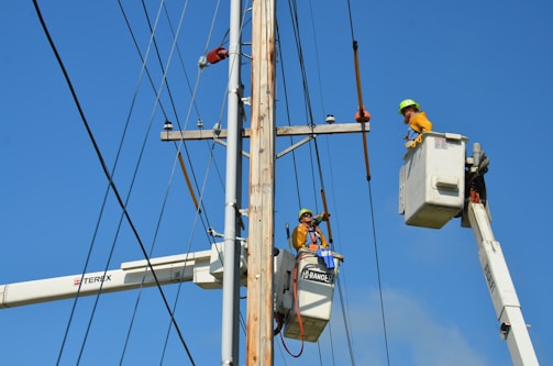 Two utility workers in safety gear are using bucket trucks to work on a power line. The sky is clear and blue, providing a backdrop to the scene. Both workers are elevated in their respective buckets, focusing on tasks related to the maintenance or repair of the electrical infrastructure.