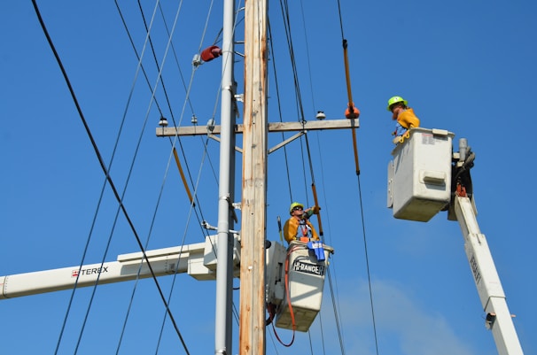 Two utility workers in safety gear are using bucket trucks to work on a power line. The sky is clear and blue, providing a backdrop to the scene. Both workers are elevated in their respective buckets, focusing on tasks related to the maintenance or repair of the electrical infrastructure.