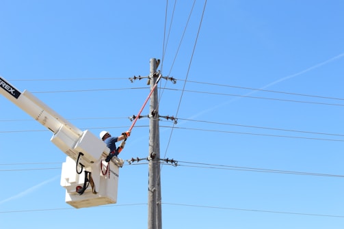 A worker in a safety harness is operating a lift bucket to repair or maintain electrical power lines. The sky is clear and bright blue, indicating a sunny day. The worker is wearing a hard hat and is holding a tool to work on the lines.