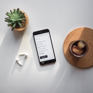 A smartphone rests on a table displaying a website. Nearby, a pair of wireless earbuds sit on the surface. A small succulent plant in a pot and a glass of iced coffee on a cork coaster complete the arrangement.