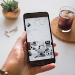 A hand holds a smartphone displaying an Instagram profile with information about a web designer and developer. On a table nearby are a pair of wireless earbuds, a small potted plant, and a glass of iced coffee on a coaster.