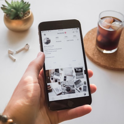 A hand holds a smartphone displaying an Instagram profile with information about a web designer and developer. On a table nearby are a pair of wireless earbuds, a small potted plant, and a glass of iced coffee on a coaster.