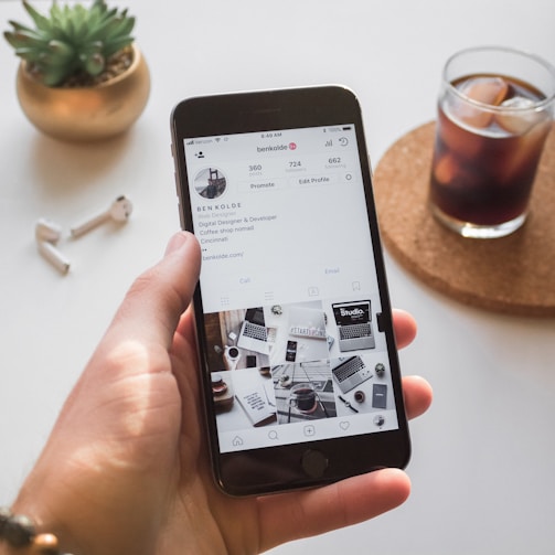A hand holds a smartphone displaying an Instagram profile with information about a web designer and developer. On a table nearby are a pair of wireless earbuds, a small potted plant, and a glass of iced coffee on a coaster.