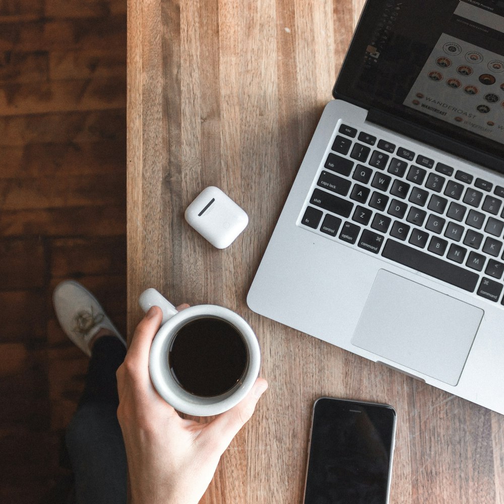 person holding cup of coffee beside gray laptop computer