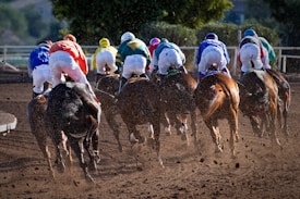 A group of jockeys riding on horses are racing on a dirt track. Their colorful uniforms create a vivid display against the backdrop of the racecourse. Dust is being kicked up by the horses' hooves, suggesting speed and movement.