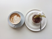 A cup of coffee with a latte art design on a white saucer placed beside a plate of dessert, possibly tiramisu, topped with cocoa powder and a decorative fruit. The dessert is served on a white plate with a small fork and a napkin.