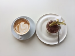 A cup of coffee with a latte art design on a white saucer placed beside a plate of dessert, possibly tiramisu, topped with cocoa powder and a decorative fruit. The dessert is served on a white plate with a small fork and a napkin.