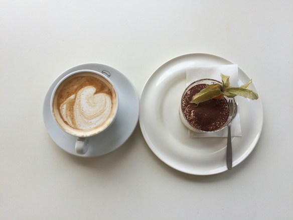 A cup of coffee with a latte art design on a white saucer placed beside a plate of dessert, possibly tiramisu, topped with cocoa powder and a decorative fruit. The dessert is served on a white plate with a small fork and a napkin.