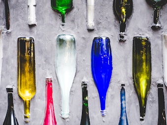 A variety of colorful glass bottles are embedded in a concrete wall, arranged vertically. The bottles are of different colors including green, yellow, blue, and red. The reflective surface of the bottles contrasts with the rough texture of the concrete.