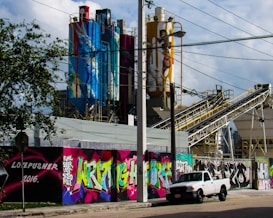 An industrial site features large silos decorated with vibrant graffiti art. A white pickup truck is parked in front, and colorful mural artwork covers the walls alongside the street. There are power lines overhead and some trees in the foreground.